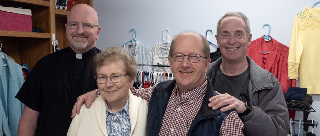 Four smiling people standing together in a clothing room, with garments hanging behind them