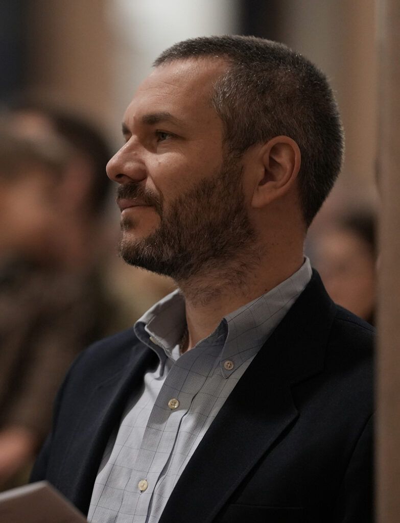 Man in a blazer and checkered shirt seated in profile at an indoor event