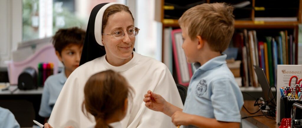 A Carmelite sister and teacher sitting at a touble with elementary-aged children, smiling at one child in particular as he talks to her.