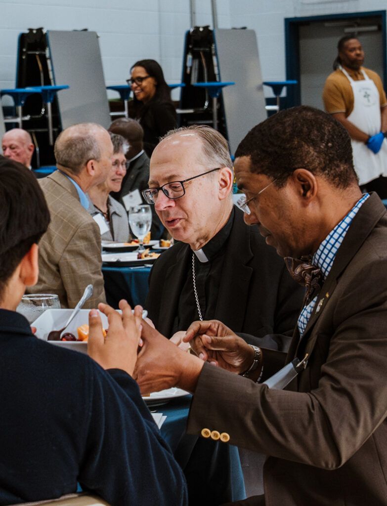 People talking over a meal at a banquet table, with a priest in the center and a man in a suit beside him